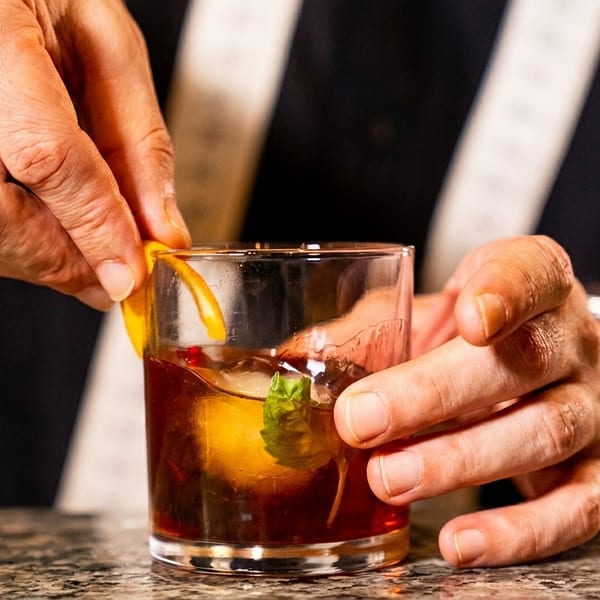 A bartender garnishes a dark cocktail with an orange peel and mint leaf on a marble bar.