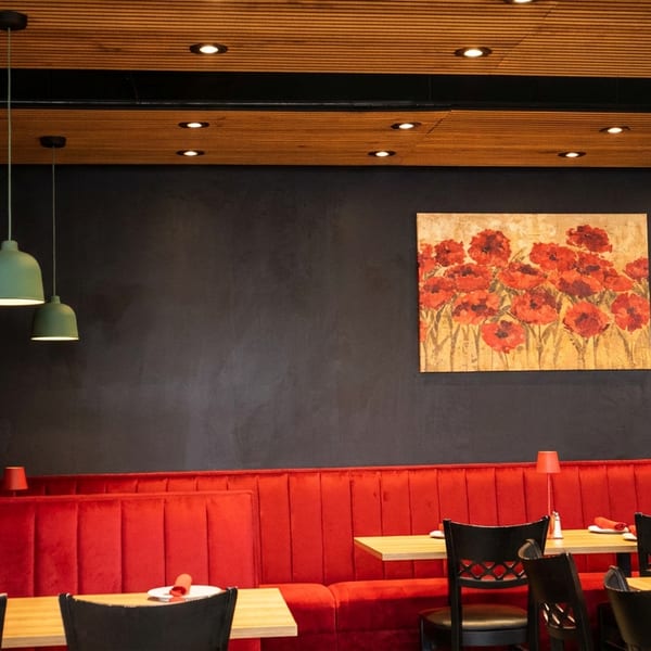 Restaurant interior with red booth seating, wooden tables, black chairs, and a poppy painting on a dark wall.