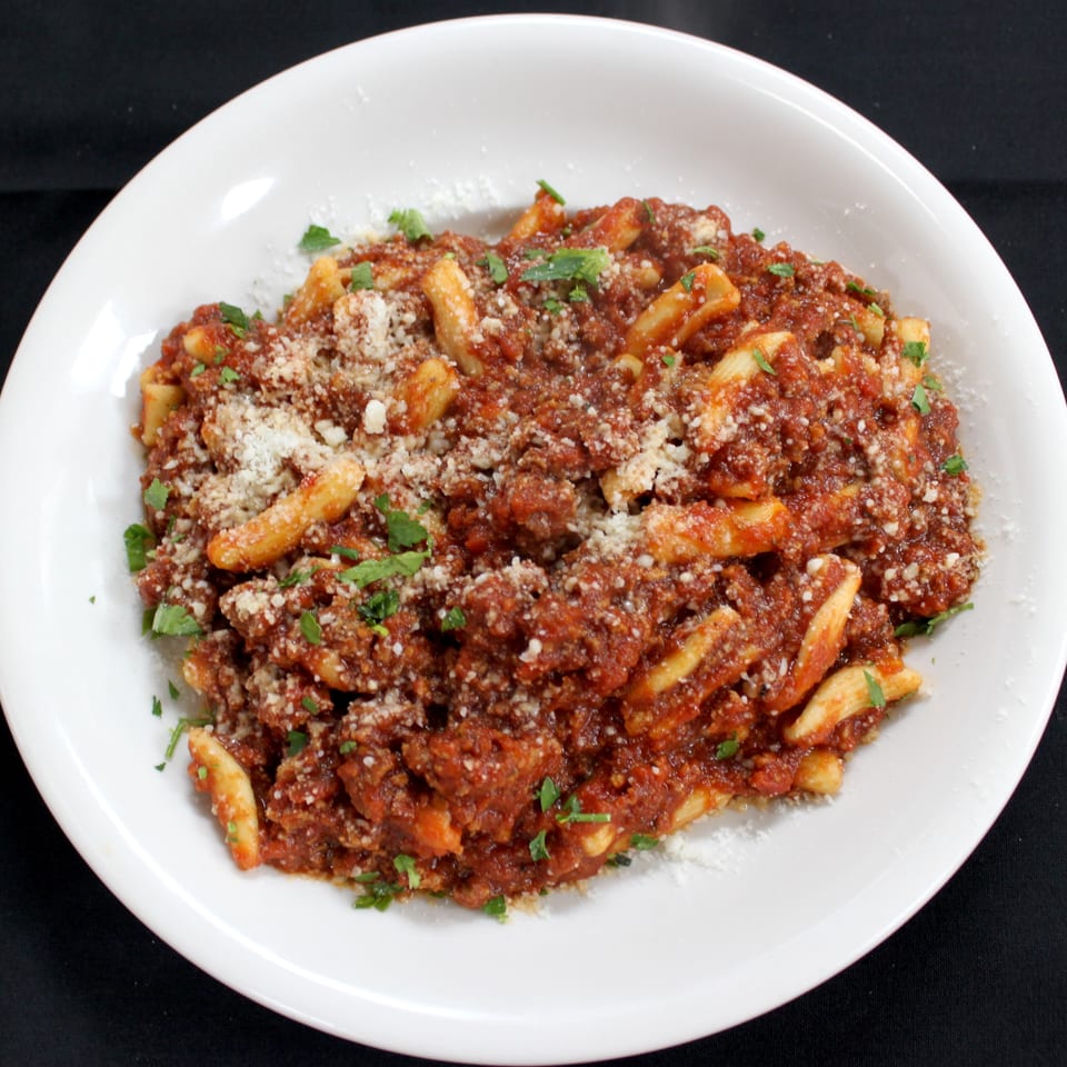 Penne pasta topped with chunky tomato meat sauce, grated cheese, and fresh parsley on a white plate.