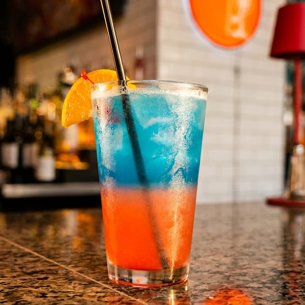 A vibrant blue and orange layered cocktail with an orange slice and straw on a bar.