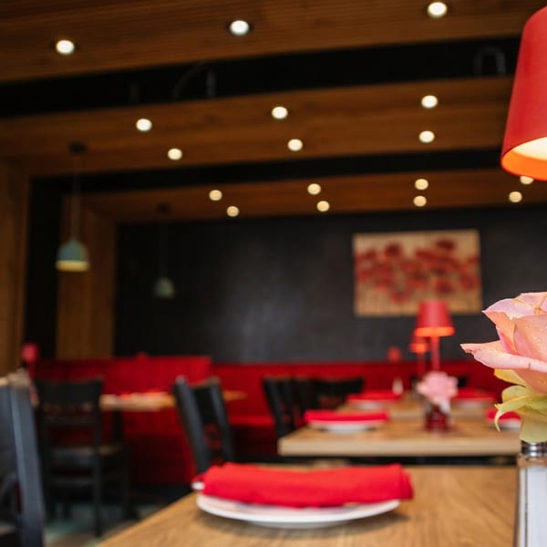 Restaurant interior with red booths, dark tables, a red lamp, and a pink rose in a vase.
