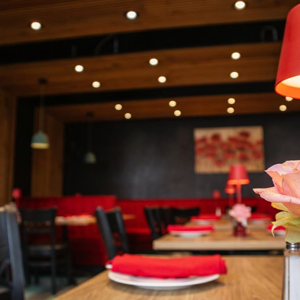 Restaurant interior with red booths, dark tables, a red lamp, and a pink rose in a vase.