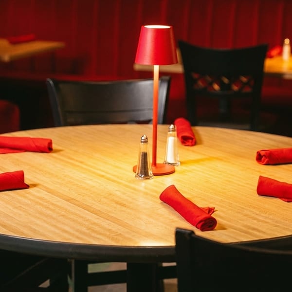 Round wooden table with red napkins, salt and pepper shakers, and a red lamp in a restaurant.