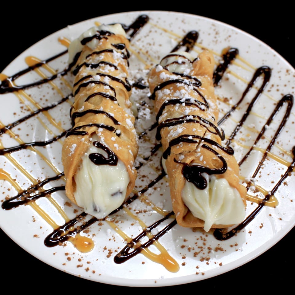 Three golden pastry cones filled with cream, drizzled with chocolate and caramel sauces, and dusted with powdered sugar.