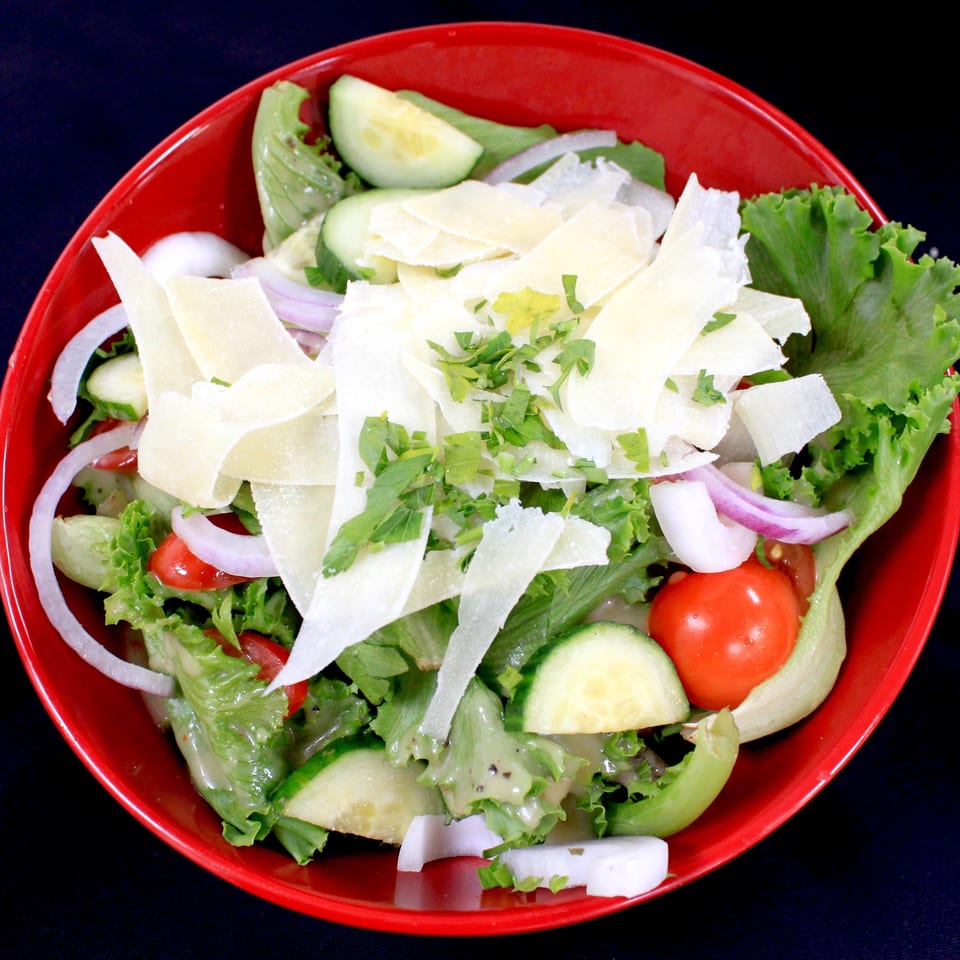 Fresh garden salad with lettuce, cucumbers, tomatoes, red onions, herbs, and shaved cheese in a red bowl.